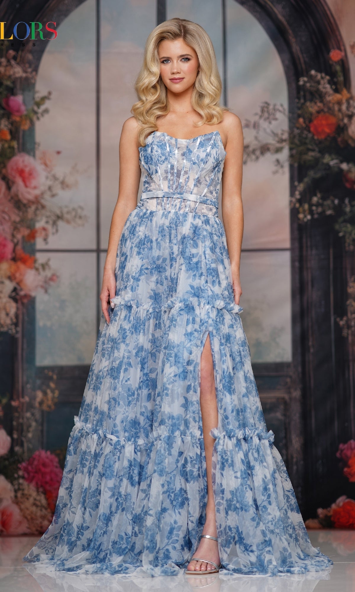 A woman models the Strapless Long Print Prom Dress: Colors Dress 3810—a strapless, floral-print gown with a high slit—standing before a floral and arch backdrop.